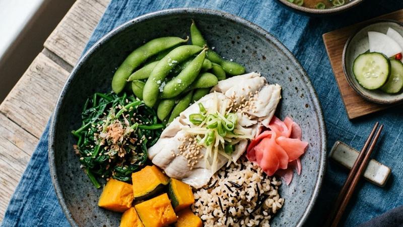A Japanese-style meal bowl with miso-glazed salmon, pickled vegetables, steamed rice, and edamame on a dark ceramic plate