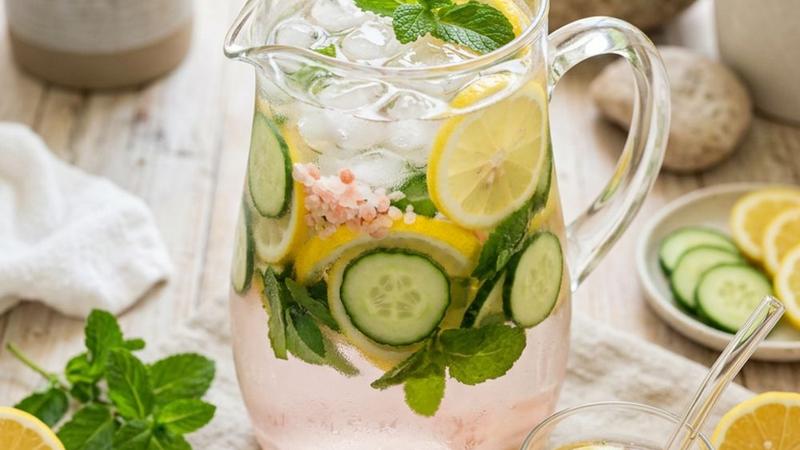 A clear glass pitcher of infused water with lemon slices, cucumber rounds, mint sprigs, and pink salt crystals