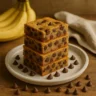 Stack of banana chocolate chip bars with chocolate chips and bananas in the background.